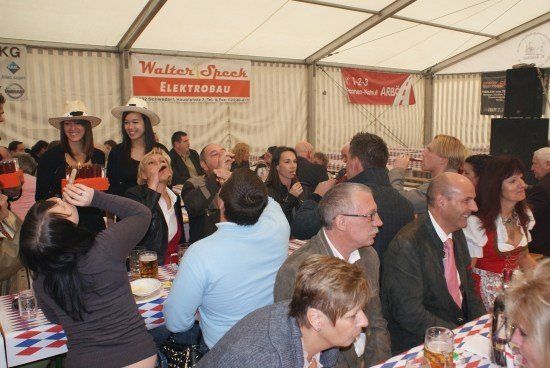 Eine Gruppe von Menschen sitzt in einem Zelt an Tischen, einige tragen Hüte und Brillen und trinken Bier. Eine Frau in einem Cowboyhut trinkt aus einem Glas.