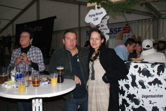 Ein Mann und eine Frau stehen lächelnd zusammen an einem Tisch mit Getränken. Dahinter zeigt ein Kuh-gemusterter Bartresen ein Schild für Jagermeister.