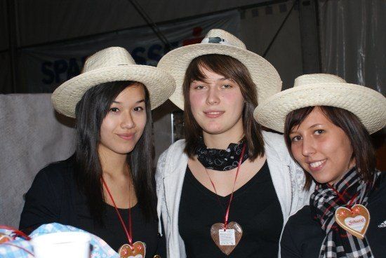 Drei Frauen mit Strohhüten und herzförmigen Namensschildern lächeln für ein Foto. Die Frau links trägt ein schwarzes Oberteil und eine rote Lanyard. Die mittlere Frau trägt eine weiße Jacke über einem schwarzen Oberteil und eine schwarze Lanyard. Die Frau rechts trägt ein schwarzes Oberteil, eine Schürze und ein herzförmiges Namensschild.