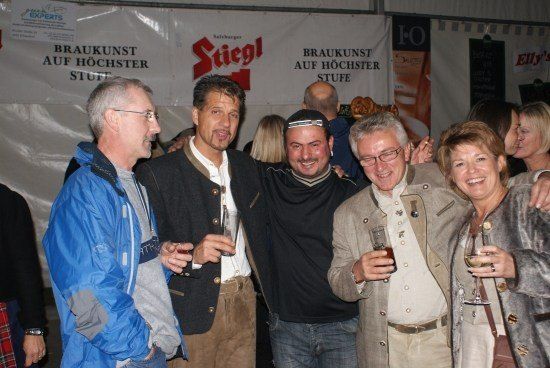 Eine Gruppe von Menschen steht bei einer Veranstaltung zusammen, lächelt und hält Gläser mit Bier in der Hand. Sie stehen vor einem Banner mit dem Wort Stiegl darauf.