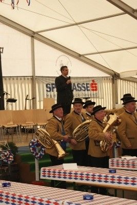 Eine Gruppe von Musikern in Uniformen spielt Blechblasinstrumente auf einer Bühne unter einem Zelt. Ein Mann in einem Anzug spricht in ein Mikrofon.