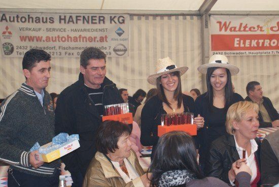 Eine Gruppe von Menschen, darunter zwei Frauen in Hüten, versammelt sich in einem Zelt, möglicherweise für eine Veranstaltung. Sie halten Boxen und lächeln. Dahinter befinden sich Banner mit Logos und Text.