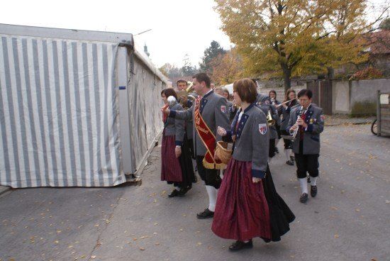 Eine Blaskapelle in traditionellen Uniformen geht eine Straße entlang, eine Frau hält einen Korb und ein Mann eine Trompete.