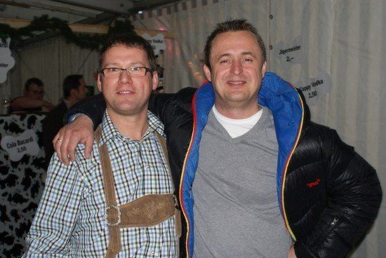 Zwei Männer lächeln und posieren für ein Foto, einer trägt ein kariertes Hemd und Hosenträger, der andere eine schwarze Jacke mit blauen Besätzen. Dahinter ein weißer Hintergrund mit Text und einem Tisch mit Kuh-Print.