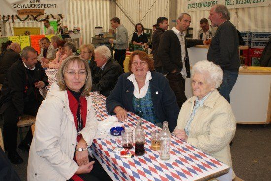 Eine Gruppe von Menschen, darunter zwei Frauen und eine ältere Frau, sitzen an einem Tisch in einer sozialen Umgebung, mit verschiedenen Getränken und Gegenständen auf dem Tisch. Im Hintergrund stehen und unterhalten sich weitere Personen.