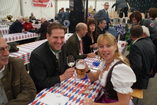 Eine Gruppe von Menschen sitzt an einem Tisch in einem Biergarten, lächelt und hebt Gläser mit Bier in die Höhe. Eine Frau in traditioneller Kleidung ist dabei. Im Hintergrund befinden sich weitere Tische und Menschen sowie eine Bühne mit Musikinstrumenten.