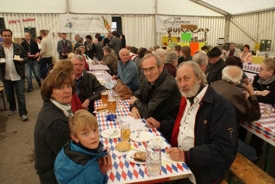 Eine Gruppe von Menschen sitzt an einem Tisch in einem Zelt, wahrscheinlich bei einem Festival. Sie genießen Essen und Getränke. Ein junger Junge in einem blauen Mantel schaut zur Kamera.