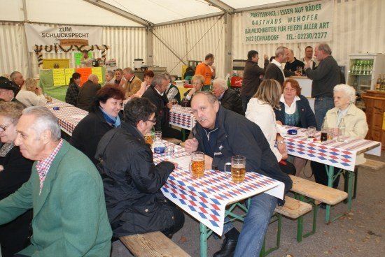 Eine Gruppe von Menschen sitzt an Tischen in einem Zelt, einige trinken Bier. Die Tische haben rot-weiß-blaue Tischdecken. Links befindet sich ein Banner mit deutschem Text.