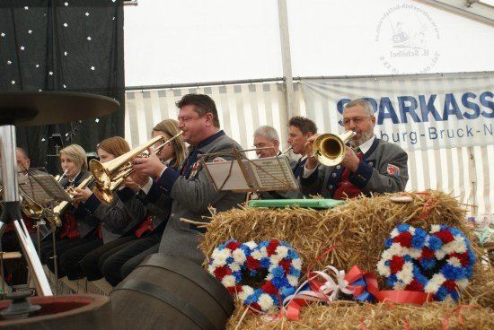 Ein Blasorchester spielt auf einer Bühne mit Heuballen und Kränzen im Vordergrund. Die Musiker tragen Uniformen und spielen Trompeten und Posaunen.