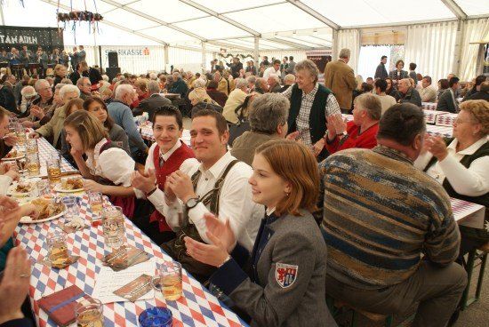 Eine große Gruppe von Menschen sitzt an langen Tischen in einem Zelt und genießt Essen und Getränke. Einige klatschen und lächeln. Die Umgebung scheint ein festliches Ereignis zu sein.