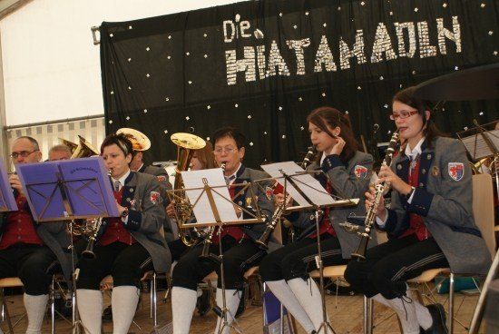 Eine Gruppe von Musikern in Uniform spielt auf der Bühne mit verschiedenen Instrumenten, darunter Trompeten und Klarinetten. Im Hintergrund steht der Text 'Die Hatamadlin'.