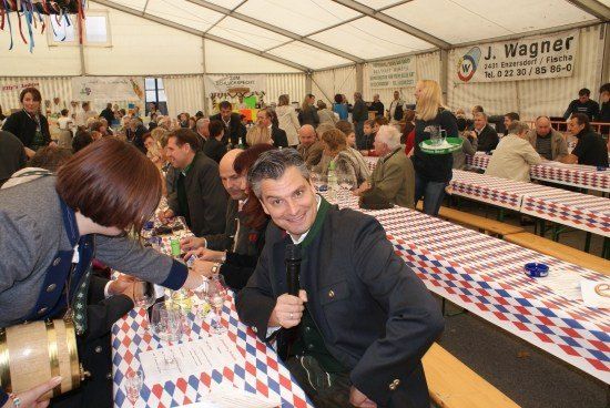 Eine Gruppe von Menschen sitzt an einem Tisch in einem Zelt, ein Mann hält ein Mikrofon und lächelt. Eine Frau schenkt Getränke in Gläser ein. Der Tisch ist mit einer rot-weiß-blau karierten Tischdecke bedeckt.