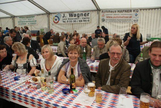 Eine Gruppe von Menschen sitzt an einem Tisch in einem Zelt und genießt Essen und Getränke. Im Hintergrund ist ein Banner mit dem Text 'J. Wagner' zu sehen.
