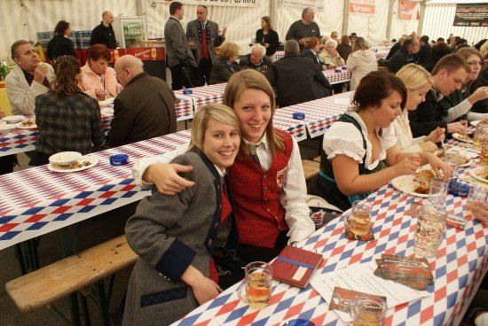 Mehrere Personen sitzen an einem langen Tisch in einem Zelt, einige in traditioneller Kleidung. Zwei Frauen lächeln und posieren für ein Foto. Der Tisch ist mit einer rot-weiß-blau karierten Tischdecke bedeckt.