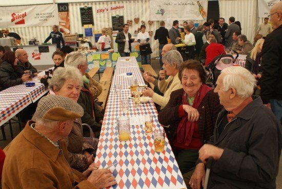 Eine Gruppe älterer Menschen sitzt an einem langen Tisch in einem Zelt, einige essen und andere trinken. Sie lächeln und scheinen sich zu amüsieren. Auf dem Tisch stehen Gläser mit Getränken und Teller.