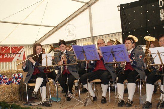 Eine Gruppe von Musikern in traditioneller Kleidung spielt auf der Bühne mit verschiedenen Instrumenten, darunter Trompeten, Flöten und Tubas. Sie sitzen auf Stühlen mit Notenständern vor sich. Die Bühne ist mit einem weißen Zelt bedeckt, und im Hintergrund befindet sich ein roter Banner mit weißem Text.