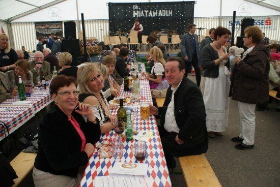 Eine Gruppe von Menschen sitzt an einem langen Tisch in einem Zelt und genießt Essen und Getränke. Der Tisch ist mit einer rot, weiß und blau karierten Tischdecke bedeckt. Hinter ihnen ist eine Bühne mit schwarzem Hintergrund aufgebaut, auf der eine Person steht.