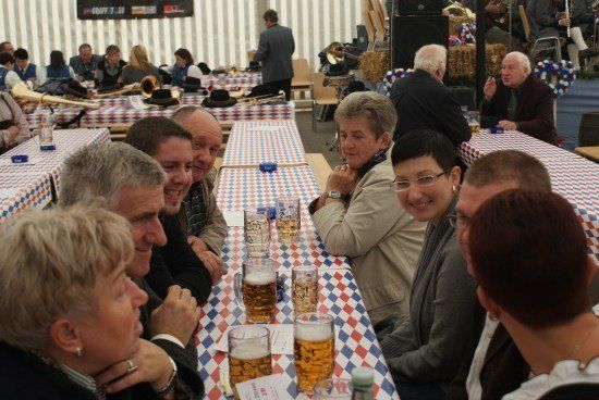 Eine Gruppe von Menschen sitzt in einem Zelt an einem Tisch, lächelt und trinkt Bier. Der Tisch ist mit einer rot-weiß-blau karierten Tischdecke bedeckt. Auf dem Tisch sind Musikinstrumente platziert. Hinter ihnen steht ein Mann in der Nähe eines Lautsprechers.