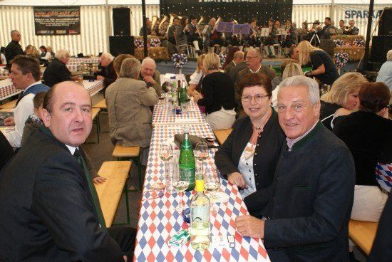 Eine Gruppe von Menschen sitzt an einem langen Tisch in einem Zelt, möglicherweise bei einem Festival. Sie lächeln und scheinen sich zu amüsieren. Der Tisch ist mit einer rot, weiß und blau karierten Tischdecke bedeckt. Auf dem Tisch stehen Flaschen, Weingläser und andere Gegenstände. Im Hintergrund spielt eine Band auf der Bühne.