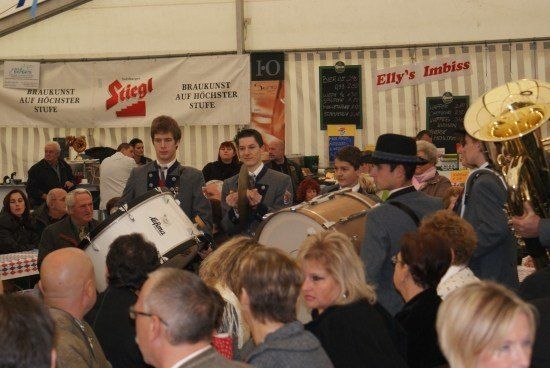Eine Gruppe von Menschen in formeller Kleidung spielt Trommeln in einem überfüllten Zelt, mit Bannern und einem Tubaspieler im Hintergrund.