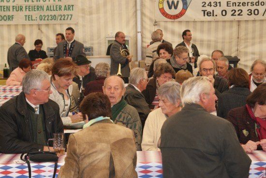 Eine Gruppe älterer Erwachsener versammelt sich in einem Zelt, einige sitzen an Tischen, während andere stehen und reden. Ein Mann hält eine französische Horn.