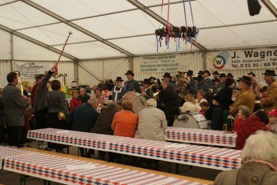 Eine Gruppe von Menschen versammelt sich in einem Zelt und beobachtet eine Marschkapelle bei der Aufführung. Die Band spielt verschiedene Instrumente, und das Publikum sitzt an Tischen mit rot-weiß-blauen Tischdecken. Das Zelt hat Banner und einen Lautsprecher.