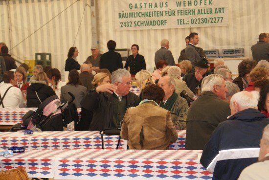 Eine Gruppe von Menschen sitzt an Tischen in einem Zelt mit einem Banner, das Gasthaus Wehofer bewirbt. Die Tische sind mit rot, weiß und blau karierten Tischdecken bedeckt.