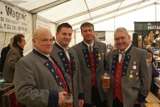 Vier Männer in traditioneller deutscher Kleidung stehen in einem Zelt, lächeln und halten Gläser mit Bier. Hinter ihnen steht ein Banner mit der Aufschrift 'Wagner' und eine Bühne ist mit einer Band aufgebaut.