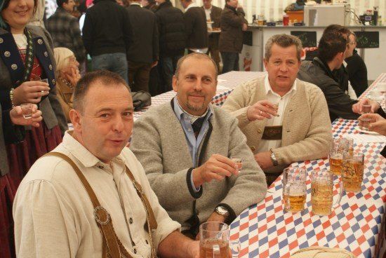 Drei Männer in traditioneller deutscher Kleidung sitzen an einem Tisch mit Bierkrügen und lächeln in die Kamera.