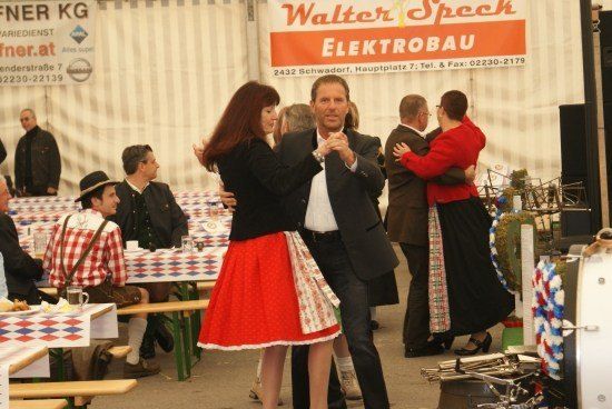Eine Gruppe von Menschen tanzt in einer festlichen Outdoor-Umgebung, mit einem Banner im Hintergrund und Tischen, die für Essen und Getränke vorbereitet sind.