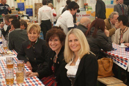 Drei Frauen in traditioneller deutscher Kleidung sitzen an einem Tisch, lächeln, mit Bierkrügen und Essen vor ihnen. Dahinter sitzen und stehen eine Gruppe von Menschen, einige in Uniform.