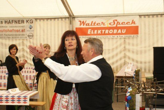 Ein Mann und eine Frau in traditioneller Kleidung tanzen in einem Zelt mit einem Banner für Walter Speck Elektrobau. Dahinter steht eine Frau in der Nähe eines Tisches mit einer farbenfrohen Tischdecke.