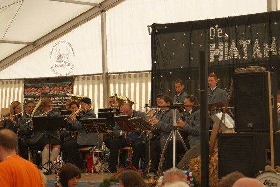 Ein Blasorchester spielt auf der Bühne in einem Zelt, mit Musikern, die verschiedene Instrumente spielen, unter einem Banner, das 'Die Phatama' steht. Das Publikum sitzt vor ihnen und beobachtet die Aufführung.
