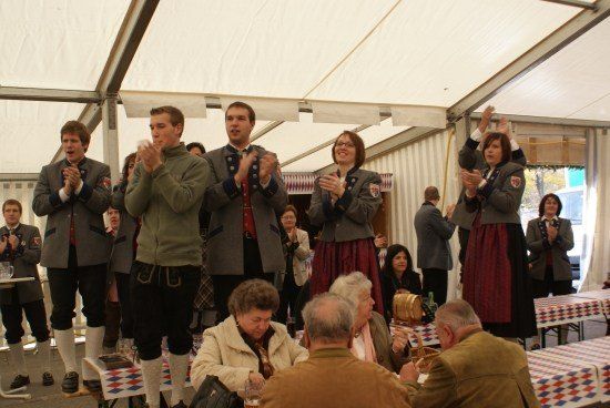 Eine Gruppe von Menschen in traditioneller Kleidung klatscht und jubelt in einem Zelt. Einige sitzen an Tischen, während andere im Hintergrund stehen.