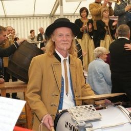 Ein Mann in traditioneller Kleidung spielt eine Trommel bei einem festlichen Ereignis, mit Musikern und Tänzern im Hintergrund.