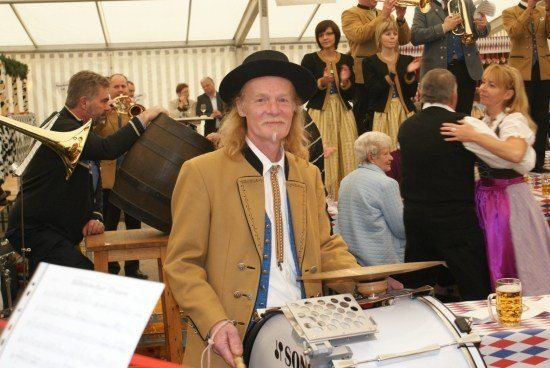 Ein Mann in traditioneller Kleidung spielt eine Trommel bei einem festlichen Ereignis, mit Musikern und Tänzern im Hintergrund.