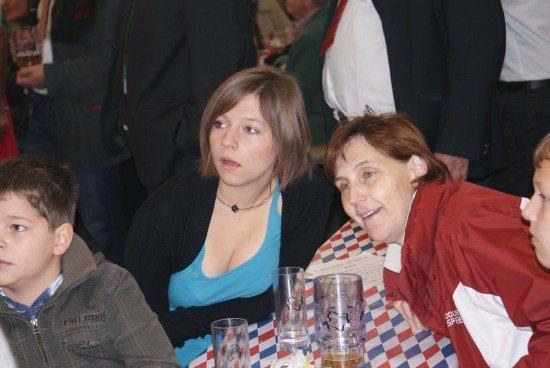 Eine Frau und ein Junge sitzen an einem Tisch mit einem rot-weiß-blau karierten Tischtuch. Die Frau trägt ein blaues Shirt und eine Halskette. Der Junge trägt eine braune Jacke. Hinter ihnen lächelt eine Frau und schaut in die Kamera. Im Hintergrund sind weitere Personen zu sehen.