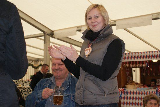 Eine Frau in einer grauen Weste klatscht mit den Händen, während sie neben einem Mann steht, der ein Glas Bier in einem festlichen Zelt hält.