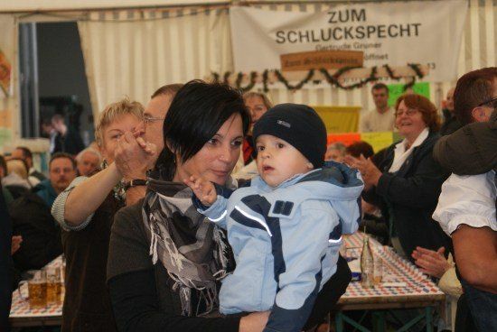Eine Frau hält ein kleines Kind in einer blauen Winterjacke bei einem festlichen Treffen, während andere im Hintergrund klatschen und lächeln.