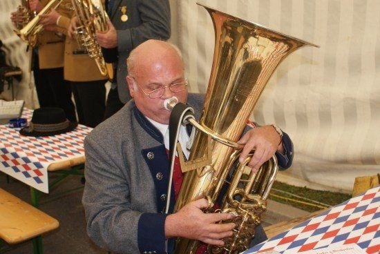 Ein älterer Mann spielt eine Messingtuba bei einer Veranstaltung im Freien, mit einem Tisch, der mit einer rot-weiß-blau karierten Tischdecke bedeckt ist, im Vordergrund.
