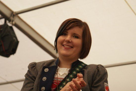 Eine Frau mit kurzen Haaren trägt ein traditionelles Kostüm mit dekorativen Knöpfen und einem roten Aufnäher am Ärmel. Sie lächelt und klatscht in die Hände.
