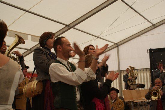 Eine Gruppe von Menschen in traditioneller Kleidung klatscht und lächelt unter einem weißen Zelt, während Musiker im Hintergrund Blechblasinstrumente spielen.