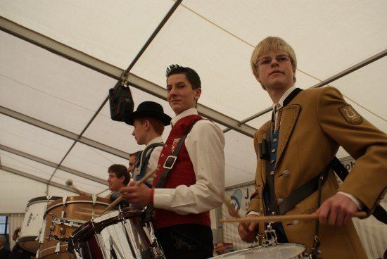 Eine Gruppe von Männern in traditioneller Kleidung spielt Trommeln unter einem Zelt. Ein Mann trägt einen schwarzen Hut.