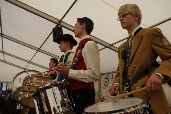 Vier Männer in traditionellen Uniformen spielen Trommeln unter einem Zelt, wobei einer eine Brille und ein anderer einen Hut trägt.