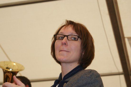 Eine Frau mit kurzen Haaren und einer Brille schaut nach oben, möglicherweise auf ein Musikinstrument. Sie trägt einen grauen Pullover und einen blauen Kragen. Hinter ihr ist ein verschwommenes Bild einer Person und einer goldenen Tuba zu sehen.