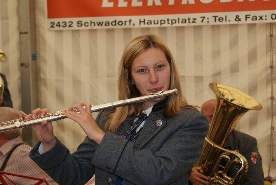 Eine Frau in einer Schuluniform spielt eine Flöte, während ein Mann hinter ihr eine Tuba spielt. Sie sind in einem Raum mit einem Banner im Hintergrund.