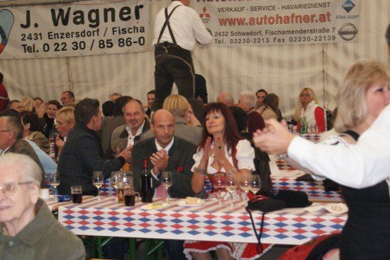 Eine Gruppe von Menschen sitzt an Tischen in einem Zelt, einige klatschen. Ein Mann steht auf einem Tisch im Hintergrund. Banner mit Kontaktinformationen sind sichtbar.
