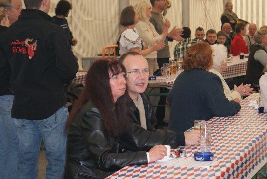 Eine Gruppe von Menschen ist in einem Zelt versammelt, einige sitzen an Tischen mit karierten Tischdecken, während andere stehen und klatschen. Ein Mann und eine Frau sitzen zusammen, lächeln und schauen zur Kamera.