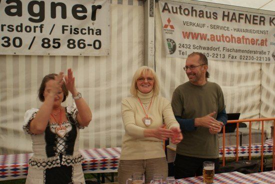 Drei Personen stehen vor einem Tisch mit Getränken, eine Frau in traditioneller bayerischer Kleidung klatscht in die Hände. Hinter ihnen werben Banner für Autohaus Hafner und Fischandorf.