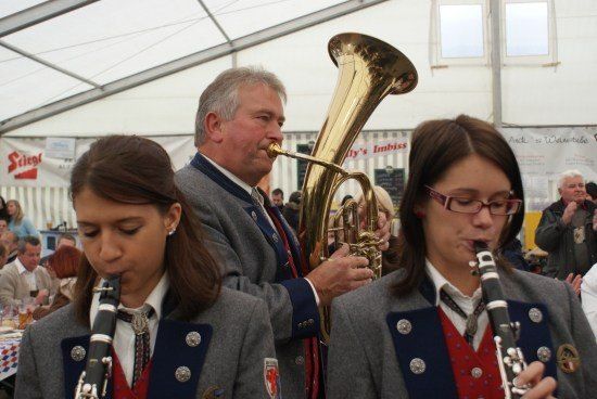 Ein Mann und zwei Frauen in gleichen Uniformen spielen Musikinstrumente, wobei der Mann ein Tuba und die Frauen Klarinetten spielen. Sie treten bei einer Veranstaltung im Freien auf.